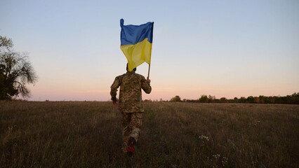 Soldier of ukrainian army running with raised blue-yellow banner on field at dusk. Young male military in uniform jogging with flag of Ukraine at meadow. Victory against russian aggression concept - 757185500