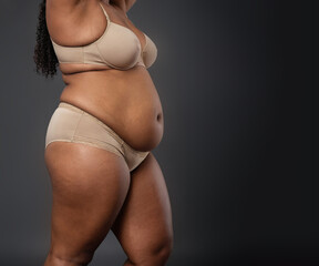 An african american dark skinned woman with a large belly is wearing a tan bra and panties. Concept of confidence and self-assurance, as the woman stands tall and proud despite her weight