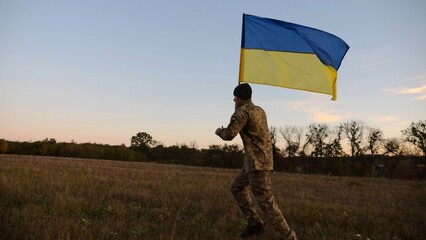 Soldier of ukrainian army running with raised blue-yellow banner on field at dusk. Young male military in uniform jogging with flag of Ukraine at meadow. Victory against russian aggression concept - 757185593