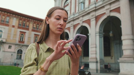 Young woman with brown hair, dressed in an olive green sweater, uses a map application on her mobile phone while standing in the old town square