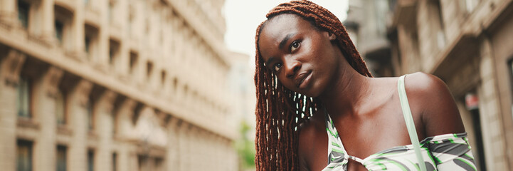 Gorgeous woman with African braids wearing top stands on the sidewalk next to the road waiting for...