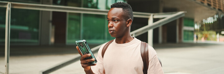 Young african student walks down the stairs outside of the university, using cellphone, Panorama