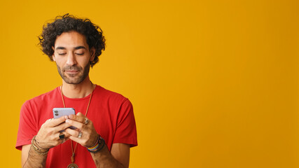 Smiling man with curly hair, dressed in red T-shirt, using mobile phone, typing message isolated on...