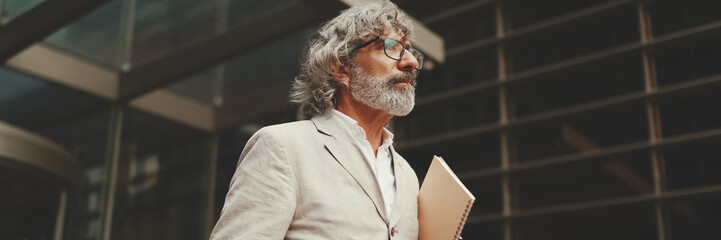 Mature businessman with beard in glasses wearing gray jacket holding folder with business documents is walking down the stairs leaving the business center, Panorama