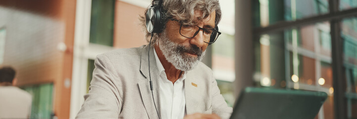 Mature businessman with beard in eyeglasses and headphones, sits in an outdoor cafe and works using...