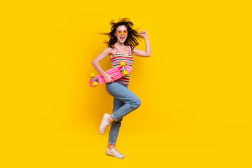 Full body photo of active girl fluttering hair wear knit tank hold skateboard run to empty space isolated on yellow color background