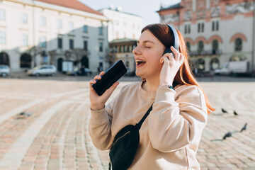 Closeup portrait of a lovely young girl listening to music through wireless earphones on urban background. Music lover enjoying music. Woman record voice by mobile cell phone dictaphone