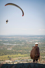 A paraglider, waiting for the right wind, watches their companion already in the air. Cityscape in...