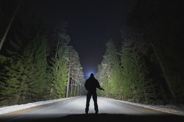 Silhouette of a man with a headlamp and a pistol in his hand on a forest road at night.