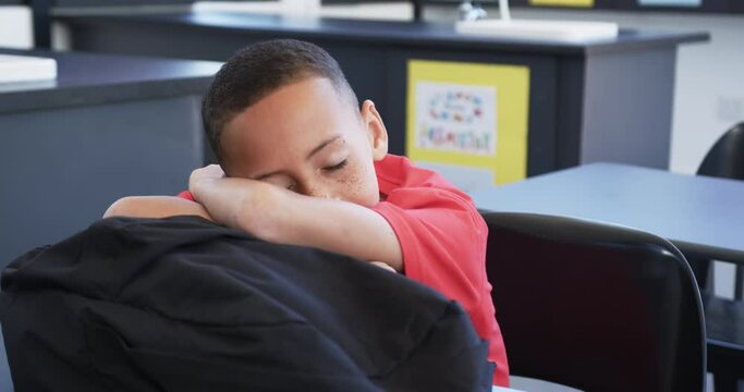 Biracial boy with freckles, wearing a red shirt, rests his head on his arms in a classroom at school
