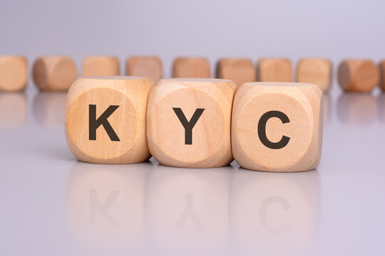 the image depicts three wooden blocks with the letters 'KYC' in focus, reflecting on the table surface. in the background, there is a row of wooden blocks, blurred and out of focus