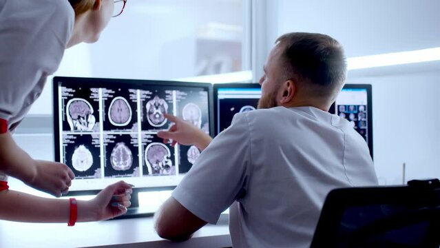In Control Room Doctor and Radiologist Discuss Diagnosis while Watching Procedure and Monitors Showing Brain Scans Results, In the Background Patient Undergoes MRI or CT Scan Procedure.