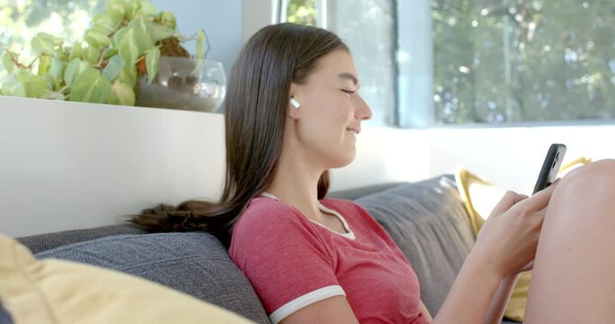 Teenage Caucasian girl with brown hair is using her smartphone, relaxing on a couch at home