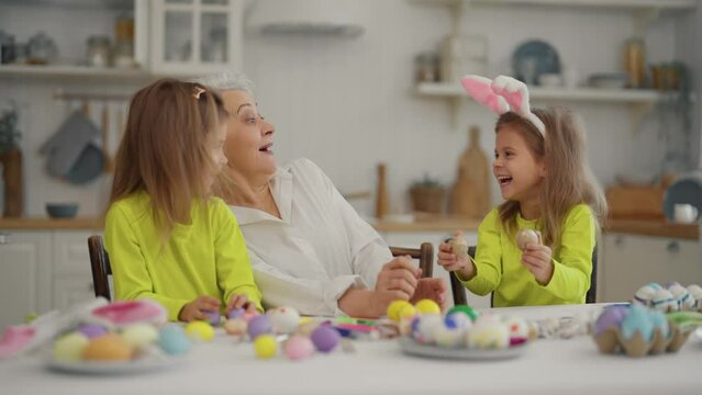 Easter grandmothers with granddaughters. Smiling grandmother with twins grandchildren wearing bunny ears decorating paint eggs together, playing with painted eggs at home. Easter leisure, have fun.