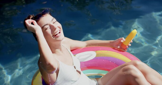 Teenage Caucasian girl enjoys the sun on a colorful float in a pool, holding a popsicle