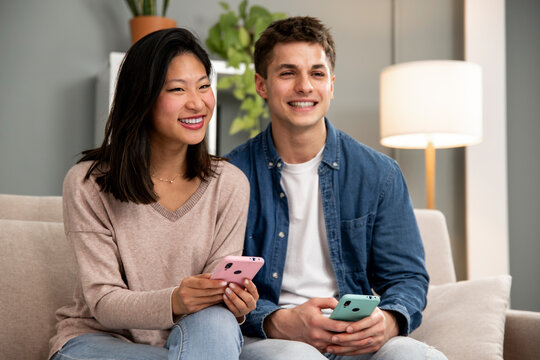 Diverse couple smiling and having fun using online phones. Joyful young adult couple using smartphone relaxing on a couch in their living room at home.