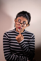 A woman with glasses is looking at the camera and pointing her finger. She is wearing a striped shirt and has a red lipstick on. Concept of curiosity and thoughtfulness