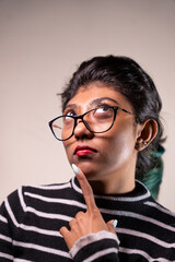 A woman with glasses is looking at the camera and pointing her finger. She has red lipstick on and her hair is pulled back. Concept of curiosity and contemplation