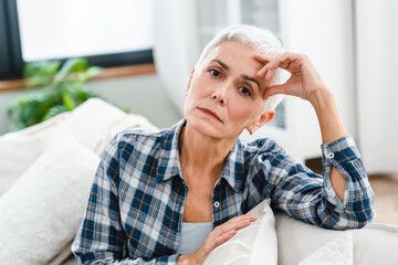 Sad woman having headache and looking at camera at home. Mature middle-aged woman sitting on the...