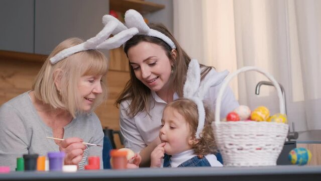 Two women and a little girl with bunny ears are happily painting Easter eggs at a table. 