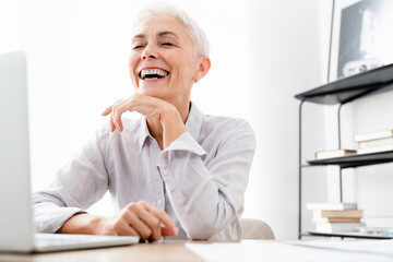 Happy Caucasian mature businesswoman winning money laughing at workplace. Middle-aged CEO manager working on laptop celebrating achievements