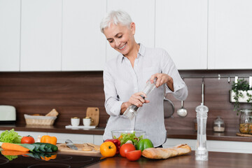 Caucasian mature housewife seasoning with spices salt pepper vegetable salad while cooking at home...