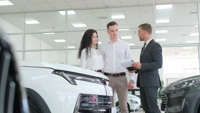A man and a woman are talking to a car dealer in a car showroom. They smile at the sleek design.