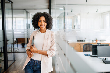 A confident young woman with a bright smile stands by a glass partition, holding the laptop in her...