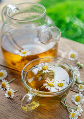 Herbal chamomile tea and chamomile flowers near teapot and tea glass on wooden table. Countryside background. - 757257527