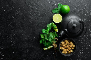 Mint tea with lime: fresh green limes, mint, brown sugar and a kettle of boiling water. On a black stone background. Top view.