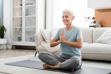 Middle-aged woman meditating in lotus position while having training at home on fitness mat. Female...