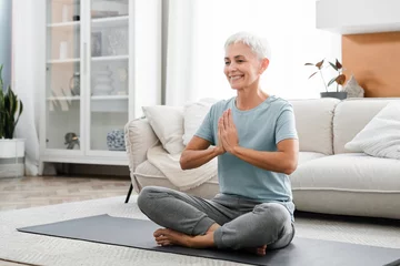 Poster Middle-aged woman meditating in lotus position while having training at home on fitness mat. Female athlete praying breathing while doing yoga indoors © InsideCreativeHouse