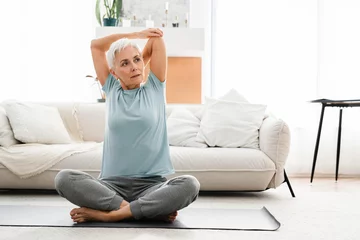 Gordijnen Fit mature woman in sporty clothes stretching while doing yoga at home on fitness mat. Middle-aged female athlete meditating in lotus position © InsideCreativeHouse