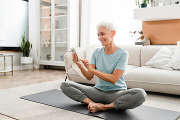Middle-aged flexible woman in fitness outfit sitting in lotus position while using cellphone for sport app. Caucasian mature woman doing online training tutorial - obrazy, fototapety, plakaty