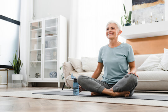Middle-aged woman doing yoga at home on fitness mat. Caucasian mature female athlete meditating in lotus position, doing training indoors on the floor