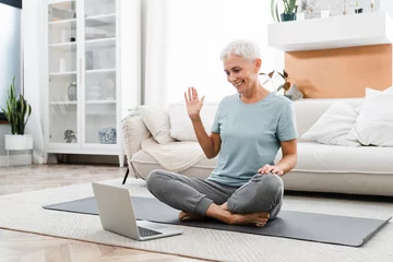 Schilderijen op glas Middle-aged woman doing sporty exercises while sitting in lotus position on videocall on laptop at home. Tutorial, online fitness class training with coach © InsideCreativeHouse