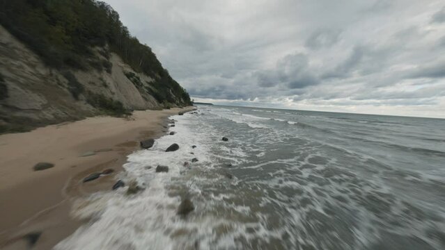 FPV drone flight above picturesque stormy foamy ocean waves washing rocky coast with cliffs under gloomy sky in overcast weather