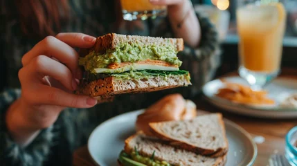 Fotobehang Close-Up of Woman's Hand Enjoying Vegetarian Sandwich with Avocado Spread © Maximilien