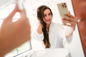 A smiling and confident Caucasian woman in a bathrobe enjoys taking selfies with her smartphone in the mirror in the bathroom with happiness to check her beautiful face and healthy hair. - 757280155