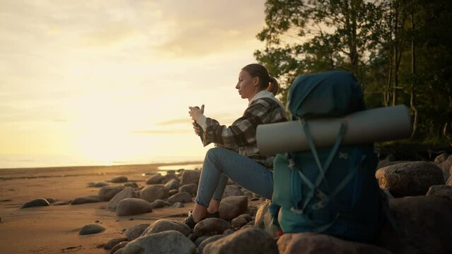 Tourist woman drinks tea on sea beach. Female having break in hike enjoying hot tea from thermos admiring nature at sunset sitting on stones. Camping hiking tourism travel wanderlust concept.