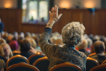 senior woman with hand up to talk at local town hall meeting, from back view