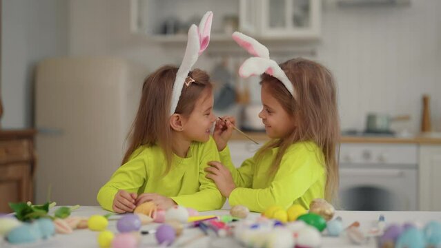 Twin girls celebrate Easter holiday. Twins cheerful children in rabbit bunny ears paint each other's faces at Easter, decorated painted colorful eggs, pastry. Easter, family having fun concept.