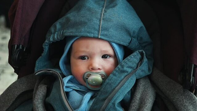 Portrait of a cute sad Caucasian baby lying in a stroller and sucking a pacifier while looking at camera