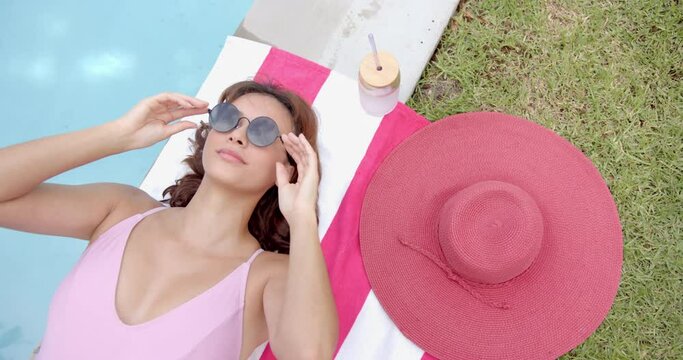 A woman in a pink swimsuit relaxes by the pool, adjusting her sunglasses
