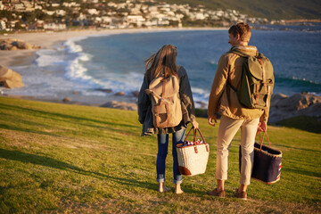 Love, walking and couple on hike with ocean, sunset for tropical holiday adventure, relax and bonding together. Picnic, man and woman on romantic date on beach, nature and grass on vacation from back