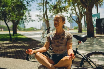 Tuinposter middle-aged latin woman meditating in lotus pose sitting on a bench next to a bicycle in a city park © juanpablo