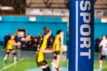 Team volleyball game, inscription SPORT on counter, selective focus. Defocused photo of volleyball court at active match in sport school gym. Sports games, team success concept. Copy ad text space