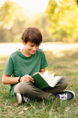 Vertical outdoors shot of a smiling teen boy sitting on grass and reading a book. © Vulp