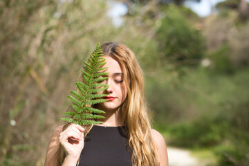 Portrait of a young, beautiful blonde woman dressed in black, half of her face is covered with a green leaf in the forest. The woman is at peace with herself.