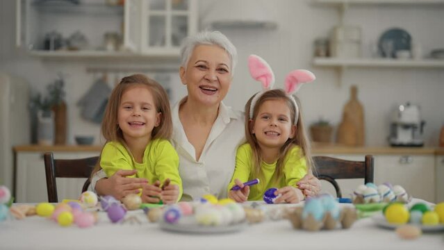 Easter grandmothers with granddaughters. Smiling grandmother hugging twins grandchildren wearing rabbit bunny ears, decorating paint eggs together in kitchen at home. Easter celebration concept.